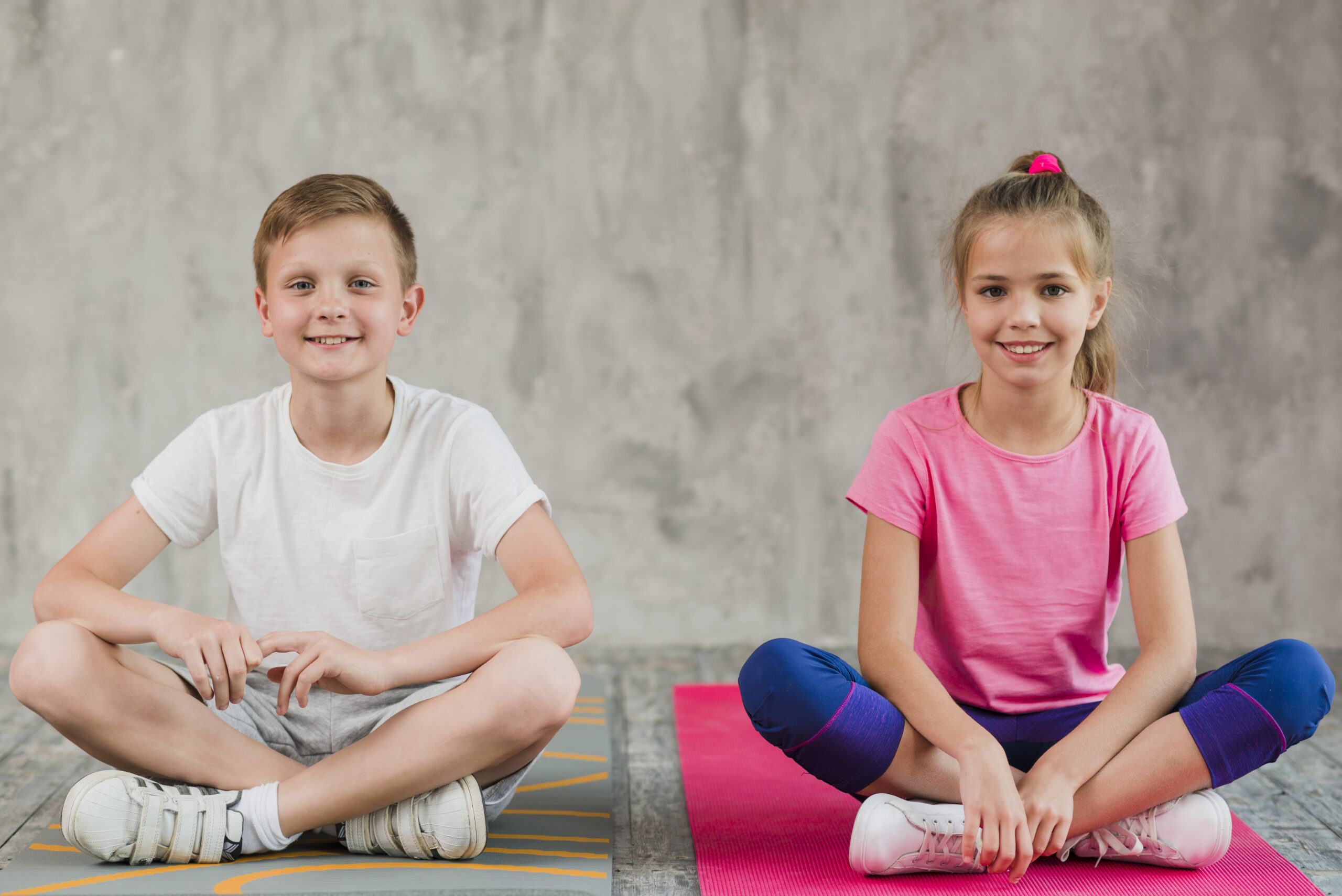 portrait-smiling-girl-boy-sitting-exercise-mat-with-their-crossed-legs-front-wall